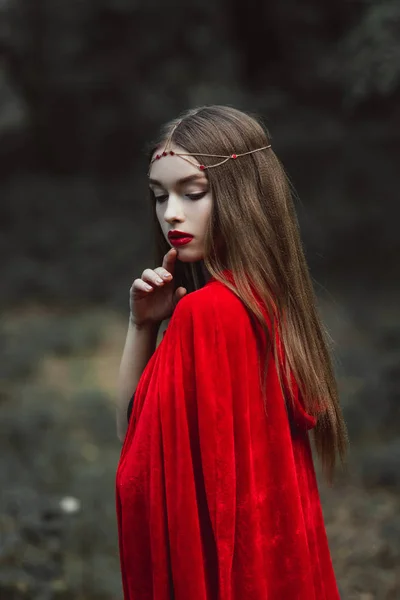 Мистическая девушка в красном плаще и элегантный венок в лесу — стоковое фото