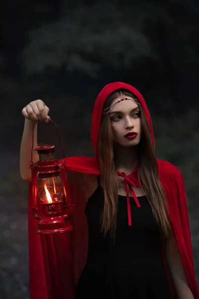 Attractive mystic girl in red cloak with kerosene lamp walking in dark forest — Stock Photo