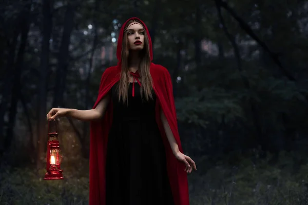 Menina mística elegante em manto vermelho com lâmpada de querosene andando em madeiras escuras — Fotografia de Stock