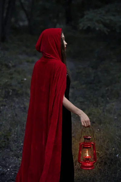 Mystic girl in red cloak walking in dark forest with kerosene lamp — Stock Photo