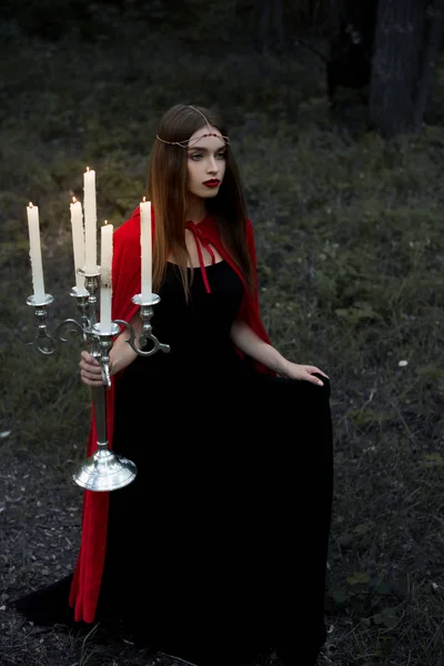 Fille mystique en manteau rouge tenant candélabre avec des bougies flamboyantes marchant dans la forêt — Photo de stock