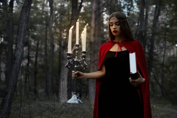 Chica mística en capa roja sosteniendo candelabro con velas encendidas y libro mágico en bosques oscuros - foto de stock