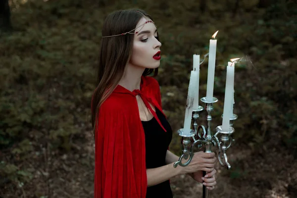 Elegant mystic girl in red cloak and wreath blowing candles in candelabrum in dark forest — Stock Photo