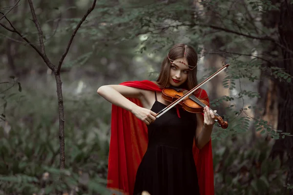 Menina mística atraente em manto vermelho tocando no violino na floresta — Fotografia de Stock