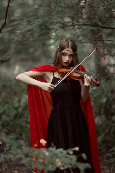 Bela menina mística em manto vermelho tocando no violino em madeiras escuras — Fotografia de Stock