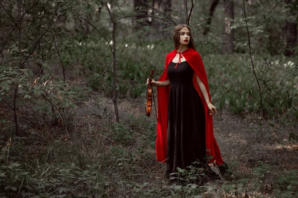 Belle fille mystique en robe noire et manteau rouge tenant violon dans la forêt — Photo de stock