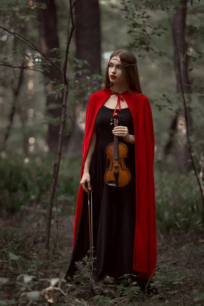 Elegante hermosa mujer en vestido negro y capa roja sosteniendo violín en el bosque - foto de stock