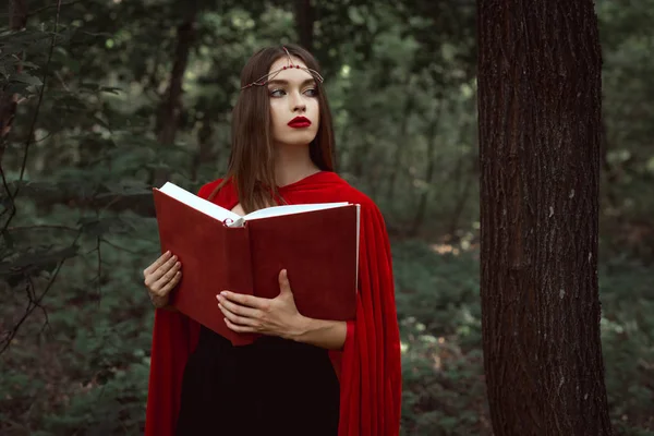 Elegant mystic girl in red cloak with magic book in forest — Stock Photo