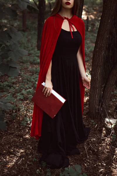 Recortado vista de chica en vestido negro y capa roja con libro de magia en el bosque - foto de stock
