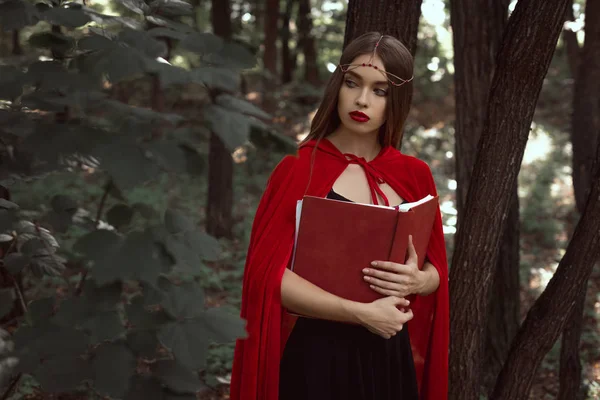 Jolie fille en manteau rouge avec livre magique dans les bois sombres — Photo de stock
