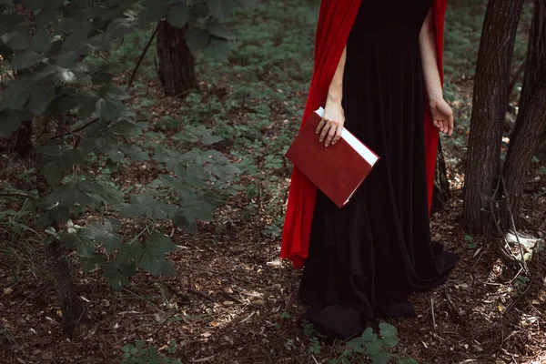 Vista recortada de chica elegante en capa roja con libro de magia en el bosque - foto de stock