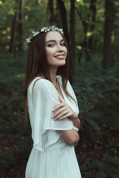 Attractive smiling girl posing in elegant dress and floral wreath in forest — Stock Photo