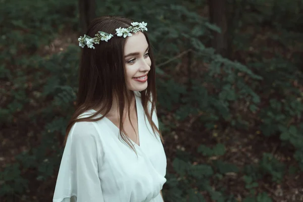 Stylish smiling girl posing in white dress and floral wreath in woods — Stock Photo