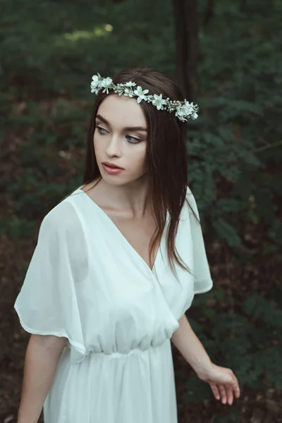 Attractive girl posing in white dress and floral wreath in forest — Stock Photo