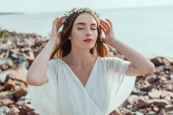 Beautiful elegant girl posing in floral wreath on rocky beach — Stock Photo