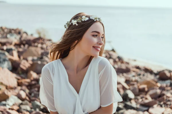 Atractiva chica sonriente posando en corona floral en la playa rocosa cerca del mar - foto de stock