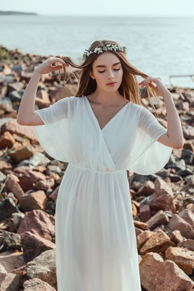 Beautiful tender girl posing in white dress and floral wreath on rocky beach — Stock Photo