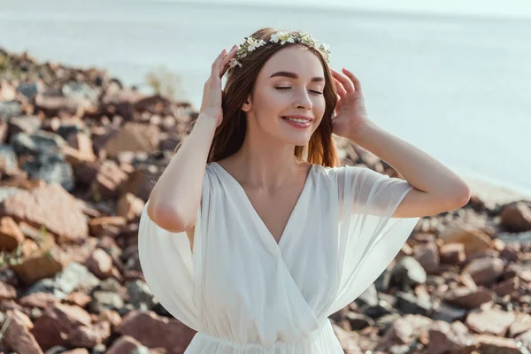 Hermosa chica sonriente posando en vestido blanco y corona floral en la playa rocosa - foto de stock