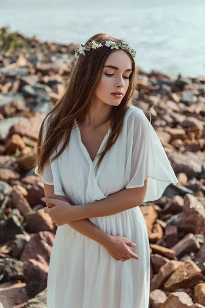 Beautiful young tender woman posing in floral wreath on rocky beach — Stock Photo