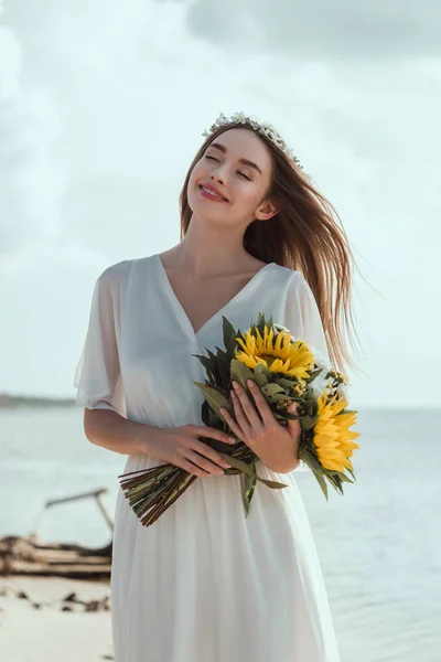 Schönes lächelndes Mädchen im weißen Kleid mit einem Strauß mit Sonnenblumen am Strand — Stockfoto