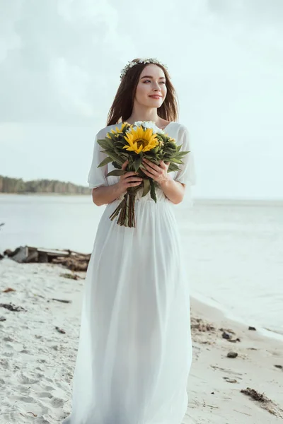 Attractive girl in elegant dress holding bouquet with sunflowers on beach — Stock Photo