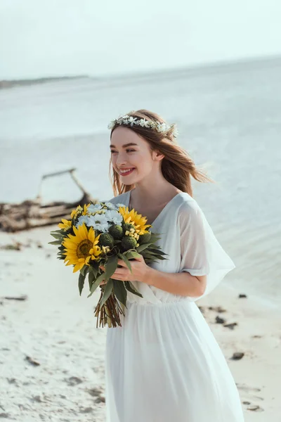 Усміхнена дівчина в білій сукні тримає букет соняшників на пляжі — стокове фото