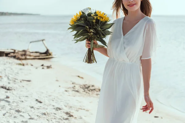 Vista ritagliata di ragazza in abito bianco che tiene bouquet con girasoli sulla spiaggia — Foto stock