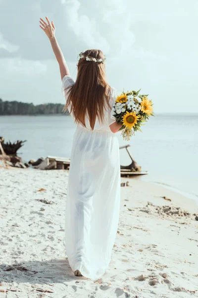 Back view of girl in white dress holding bouquet with sunflowers on seashore — Stock Photo