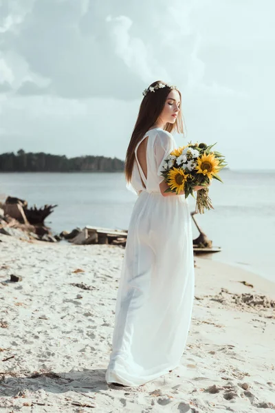 Menina elegante em vestido branco e coroa floral segurando flores na costa do mar — Fotografia de Stock