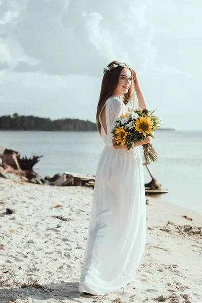 Jolie fille en robe élégante et couronne florale tenant tournesols sur le bord de la mer — Photo de stock