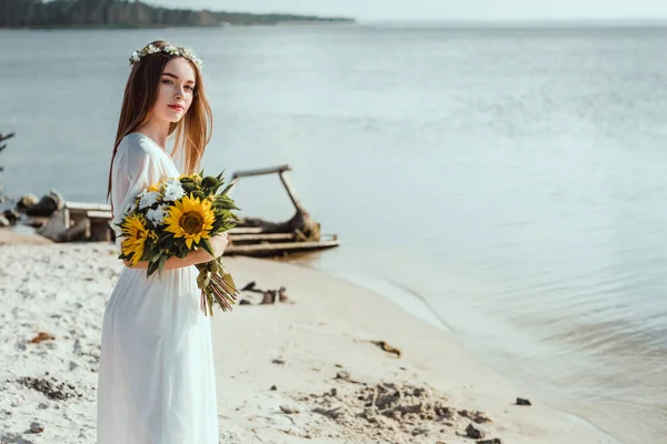 Beautiful girl in elegant dress and floral wreath holding bouquet on beach — Stock Photo