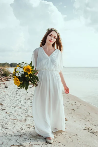Menina elegante em vestido branco segurando buquê e andando na praia — Fotografia de Stock