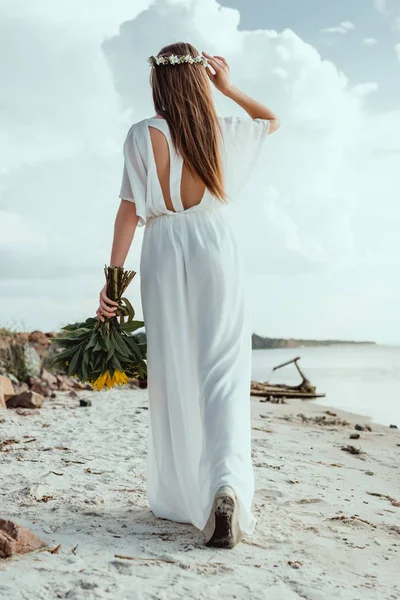 Back view of elegant girl in white dress holding bouquet on seashore — Stock Photo