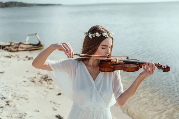 Jolie jeune femme en robe élégante et couronne florale jouant du violon sur le bord de la mer — Photo de stock