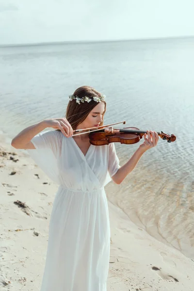 Belle femme en robe blanche élégante et couronne florale jouant du violon sur le bord de la mer — Photo de stock