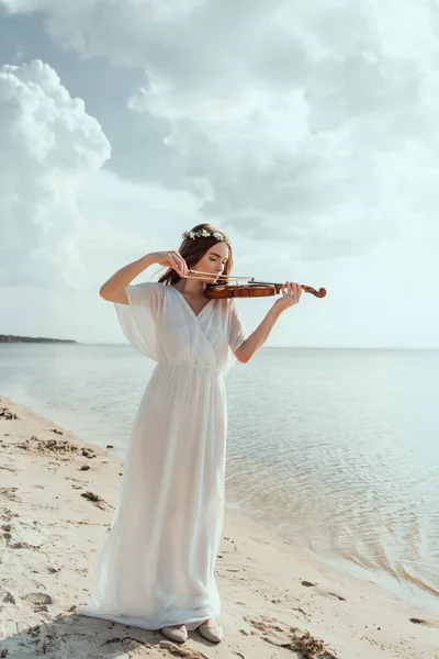 Красивая элегантная девушка в белом платье и цветочный венок играет на скрипке на песчаном пляже — стоковое фото