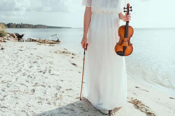 Vista recortada de chica en vestido elegante sosteniendo violín en la playa cerca del mar - foto de stock