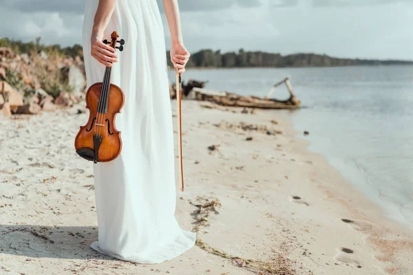 Vista cortada de menina elegante em vestido branco segurando violino na praia de areia — Fotografia de Stock