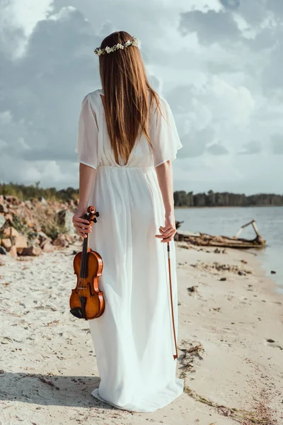 Back view of girl in elegant white dress holding violin on seashore — Stock Photo