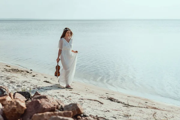 Jolie jeune femme en robe élégante avec violon marchant sur la plage près de la mer — Photo de stock