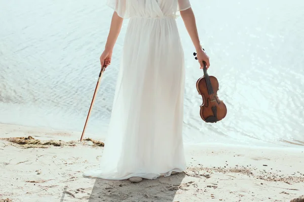 Vue recadrée de fille en robe blanche tenant violon sur le bord de la mer — Photo de stock