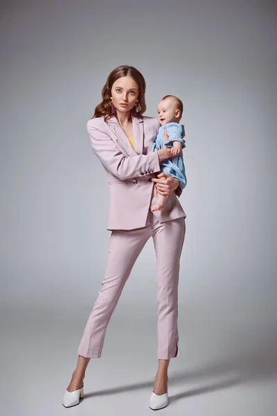 Full length view of stylish young mother holding adorable infant daughter and looking at camera on grey — Stock Photo