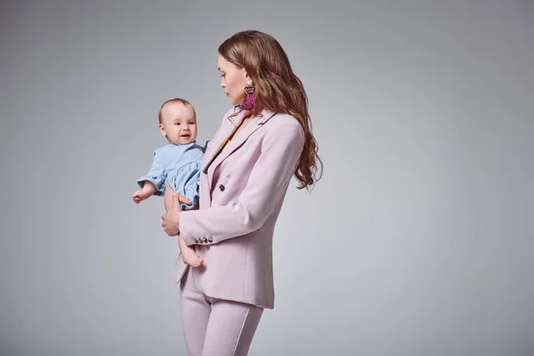 Вид збоку молодої матері в стильному рожевому костюмі, що носить чарівну доньку-немовля ізольовану на сірому — стокове фото