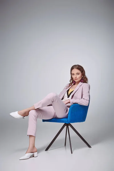 Femme élégante confiante assise sur une chaise bleue et regardant la caméra sur le gris — Photo de stock