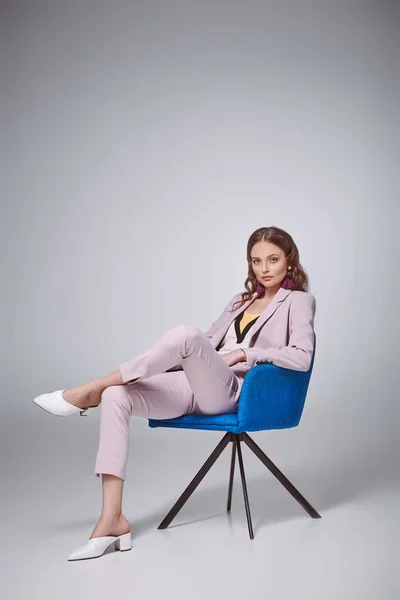 Mujer con estilo de confianza sentado en la silla azul y mirando a la cámara en gris - foto de stock