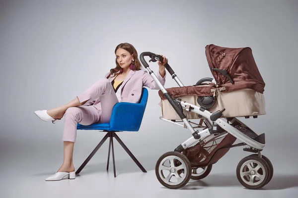Stylish woman holding baby stroller while sitting on chair and looking away on grey — Stock Photo