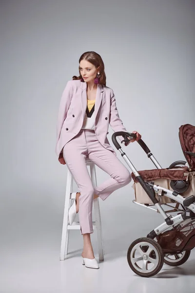 Beautiful stylish woman with baby stroller sitting on stool and looking away on grey — Stock Photo