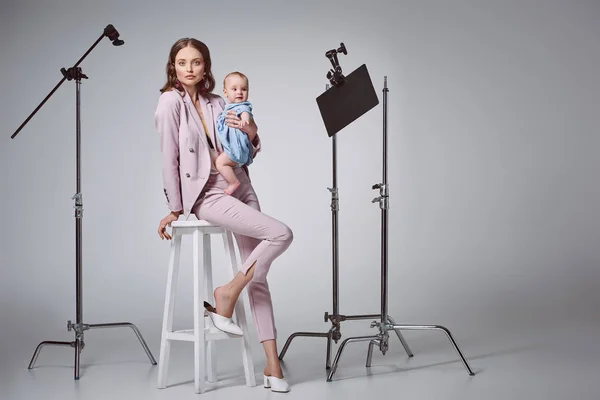 Belle mère élégante tenant adorable petite fille et regardant la caméra tout en étant assis sur le tabouret dans le studio d'enregistrement — Photo de stock