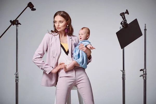 Moda joven madre holding adorable bebé hija mientras sentado en silla en estudio de grabación - foto de stock
