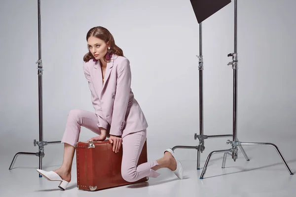 Belle femme élégante en costume rose assise sur la valise et regardant loin dans le studio d'enregistrement — Photo de stock